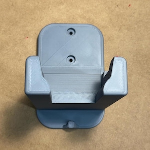 May include: A pale grey 3D-printed wall mount with a curved base and a rectangular opening. The mount has two circular holes at the top for mounting and two side supports to hold an item. The item is designed to be mounted on a wall.