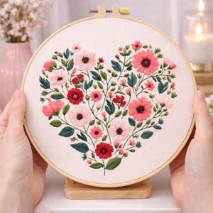 Könnte beinhalten: Ein besticktes Herzmuster mit rosa und roten Blumen und grünen Blättern auf cremefarbenem Stoff. Der Stoff ist in einem Holzrahmen gespannt und wird von einem Holzständer gehalten.