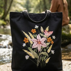 Puede incluir: Bolso tote negro con diseño floral bordado. El bordado presenta un ramo de lirios rosas, flores naranjas y blancas y detalles amarillos. El bolso tiene dos asas y está hecho de una tela resistente.