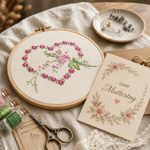 Può includere: Un telaio da ricamo con un motivo floreale a forma di cuore in rosa e verde. Un biglietto di auguri con la scritta "zum Muttertag" e un bordo floreale è accanto al telaio. Altri oggetti includono forbici, filo e un piattino.