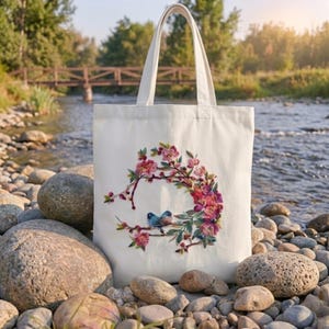 Pode incluir: Uma sacola tote creme com um design floral e de pássaros. O design apresenta flores rosa e vermelhas com dois pássaros azuis empoleirados em um galho. A sacola tem duas alças e é feita de um tecido resistente.