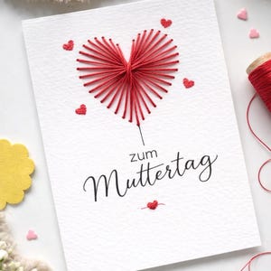 Pode incluir: Um cartão branco com um design de coração vermelho costurado e o texto alemão "zum Muttertag". O cartão apresenta pequenos corações vermelhos bordados e é acompanhado por uma bobina de linha vermelha.
