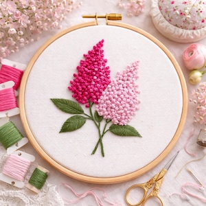 May include: An embroidery hoop with a floral design. The design features two flower clusters in shades of pink, with green leaves and stems. The embroidery is set against a white fabric, and the hoop is made of wood. Spools of thread and other sewing supplies are in the background.