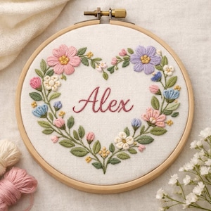 Könnte beinhalten: Ein floraler Stickrahmen mit herzförmigem Kranz aus rosa, blauen und lila Blumen mit grünen Blättern. Der Name "Alex" ist in Schreibschrift innerhalb des Herzens gestickt. Der Rahmen ist aus Holz.