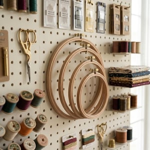 May include: A craft room display featuring wooden embroidery hoops of varying sizes, gold scissors, spools of thread in various colors, and packages of needles. The items are arranged on a pegboard, creating an organized and visually appealing workspace.
