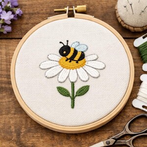 Könnte beinhalten: Gesticktes Bienen- und Gänseblümchen-Design in einem hölzernen Stickrahmen. Die Biene ist schwarz und gelb mit hellblauen Flügeln, die auf einer weißen Gänseblümchen mit gelber Mitte und grünem Stiel sitzt. Der Rahmen befindet sich auf einem Holzgrund.