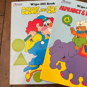 May include: Two vintage "Wipe-Off Book" educational workbooks. One book is titled "Colors and Shapes" with a clown illustration. The other book is titled "Alphabet & Numbers" with a bear riding an elephant. Both books have a light blue cover.
