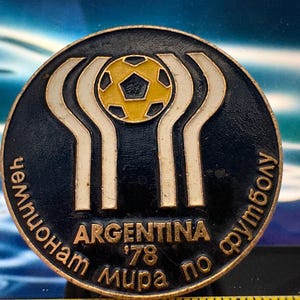 May include: A circular, dark blue metal pin featuring a yellow soccer ball at the center. The pin has a white and gold design with the text "ARGENTINA '78" and Cyrillic text around the edge. A vintage collectible.