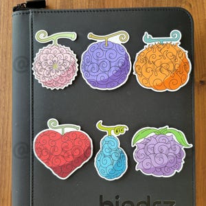 May include: A collection of six colorful fruit stickers with unique designs, including a pink floral, a purple sphere, and an orange flame-like shape. The stickers are on a black binder with the word "bindrz" embossed on it.