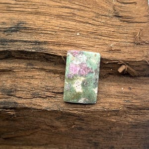 May include: A rectangular cabochon gemstone featuring a mottled pattern of deep magenta and forest green mineral inclusions. The stone has a smooth, polished surface with visible colour variations throughout, displaying a mix of ruby and zoisite.