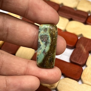 Könnte beinhalten: Ein rechteckiger, polierter Edelstein-Cabochon mit einer gesprenkelten Oberfläche in tiefem Waldgrün, erdigem Braun und kleinen dunkelmagentafarbenen Flecken. Der Stein hat eine glatte, glänzende Oberfläche und eine flache Form.