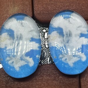 Könnte beinhalten: Zwei ovale Glaskabochons mit einem aufgedruckten Motiv aus weißen Wolken vor einem hellblauen Himmel. Die glatten, gewölbten Oberflächen haben ein glänzendes Finish, das das Licht reflektiert und jedem einzelnen Stück ein klares, abgerundetes Aussehen verleiht.