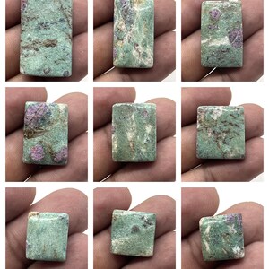 May include: Nine rectangular gemstones with a mottled appearance. The stones have a base colour of light green with patches of darker green, brown, and pinkish-red. The stones are held in a person's hand.