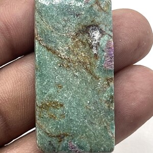 May include: A rectangular gemstone with a mottled appearance. The stone features a base color of teal with brown and purple veining. The stone is held in a hand.