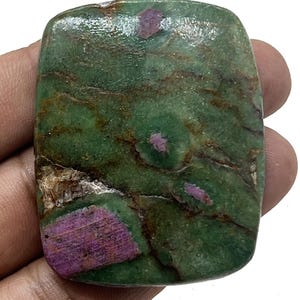 Puede incluir: Una gema rectangular con una superficie pulida. La piedra presenta un color base verde moteado con parches de rojo intenso y vetas marrones. La gema se sostiene en una mano.