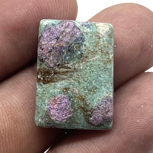 May include: A rectangular gemstone with a mottled appearance. The stone has a light green base colour with patches of purple and brown. The gemstone is held between two fingers.