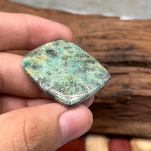 Op de afbeelding: Een rechthoekige, gepolijste edelsteen cabochon met een gevlekt oppervlak in tinten bosgroen, donker houtskool en subtiele gouden spikkels. De steen heeft een glad, licht gewelfd bovenoppervlak en afgeronde randen met een natuurlijk mineraalpatroon.