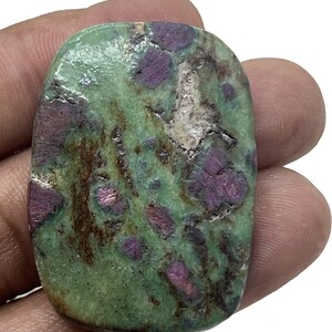 Puede incluir: Una piedra preciosa rectangular pulida, con un color base verde, salpicada de inclusiones rojo oscuro y marrón. La piedra tiene una superficie lisa, ligeramente redondeada, que muestra patrones y variaciones naturales. La gema se sostiene en una mano.