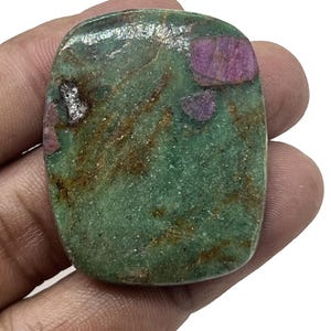 Puede incluir: Una piedra preciosa rectangular con un color base verde y parches de rojo intenso y marrón. La piedra tiene un aspecto ligeramente pulido y se sostiene en una mano.