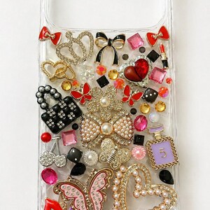 May include: Clear phone case adorned with an array of decorative charms. Features include bows, hearts, butterflies, lipsticks, and perfume bottles. Embellishments are in various colors, including red, pink, black, and gold, creating a glamorous aesthetic.