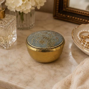 Peut inclure: Une boîte à bijoux ronde en laiton avec un couvercle amovible. Le couvercle présente une finition en émail bleu clair texturé avec un motif floral et arboré doré en relief. La base du récipient est en laiton poli avec un extérieur lisse et arrondi.