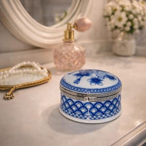 May include: A small, round porcelain trinket box with a hinged lid. The box is white with blue floral and geometric patterns. A silver-coloured metal band encircles the box. The box is on a white surface, with a mirror, perfume bottle, and flowers in the background.