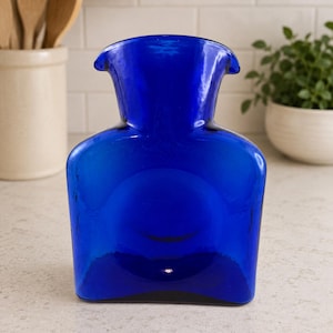 Pode incluir: Uma jarra de vidro azul cobalto vibrante com uma borda larga e alargada e um corpo liso e arredondado. O recipiente apresenta um acabamento translúcido que capta a luz, exibindo um tom azul profundo e saturado em toda a sua estrutura.
