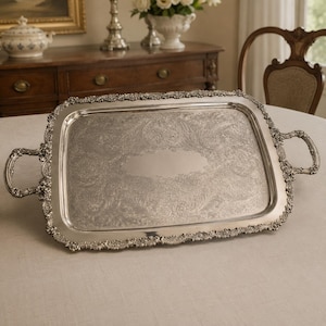 May include: A rectangular silver serving tray featuring ornate, raised floral and scrollwork detailing along the edges and handles. The centre of the tray has a smooth, polished oval area surrounded by an intricate, etched pattern of leaves and vines.