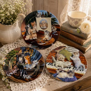 May include: Three round decorative ceramic plates featuring detailed illustrations of kittens. One plate shows kittens painting, another depicts kittens on a rocking chair, and the third displays kittens playing on a piano with sheet music.