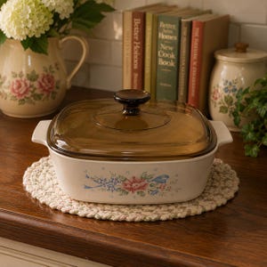 May include: A white casserole dish with a brown glass lid, decorated with a floral pattern in pink and blue. It rests on a cream-coloured, braided trivet, ideal for kitchen use. The dish is likely to be around 20 cm in length.
