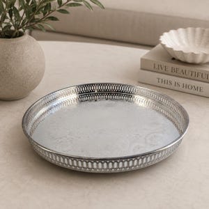 Peut inclure: Un plateau rond en métal argenté présentant un motif floral en relief complexe sur la base et un bord ajouré décoratif avec un design à fentes verticales répétitives, offrant une surface polie et réfléchissante pour la présentation.