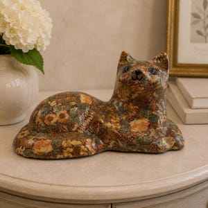 Op de afbeelding: Een decoratief kattenbeeldje met een dicht, veelkleurig bloemenpatroon in tinten oranje, geel, groen en bruin. De kat is afgebeeld in een liggende houding met de kop licht omhoog gedraaid, voorzien van donkere, amandelvormige ogen.