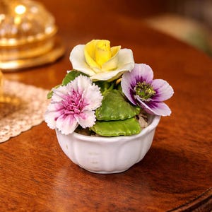 Puede incluir: Una pequeña maceta de cerámica blanca llena de un arreglo floral en miniatura. El arreglo presenta una rosa amarilla, un clavel rosa y blanco y una flor púrpura, con hojas verdes. La maceta está sobre una superficie de madera.