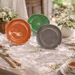 Peut inclure: Trois assiettes en céramique orange, verte et grise, chacune présentée sur un support transparent. Les assiettes ont une forme légèrement irrégulière. La scène est sur une table avec une composition florale et des couverts anciens.