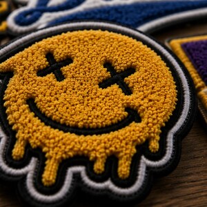 Può includere: Una toppa circolare gialla a forma di faccina sorridente con una superficie testurizzata e trapuntata. Il viso presenta occhi a forma di X cuciti in nero e una bocca larga e curva. Il bordo inferiore del viso giallo sembra sciogliersi, con gocce che si estendono verso il basso su un bordo nero e bianco.