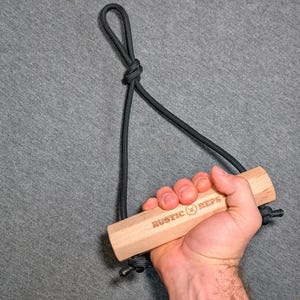 May include: A wooden exercise handle with the text "RUSTIC REPS" is held by a hand. The handle is attached to a black rope, forming a loop at the top. The handle is made of light-colored wood.
