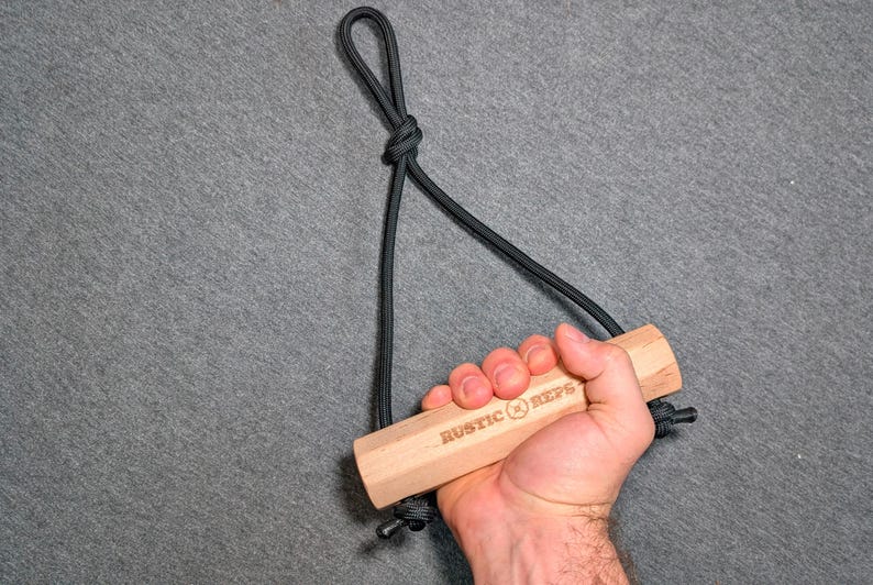 May include: A wooden exercise handle with the text "RUSTIC REPS" is held by a hand. The handle is attached to a black rope, forming a loop at the top. The handle is made of light-colored wood.