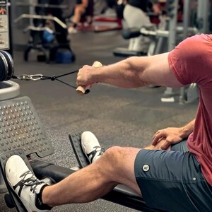 May include: A person using a seated cable row machine in a gym. The person is wearing a red t-shirt, dark blue shorts, and white sneakers. The machine has a black seat, footrests, and a cable with wooden handles.