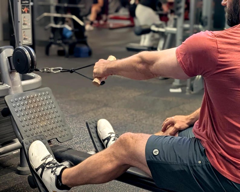 May include: A person using a seated cable row machine in a gym. The person is wearing a red t-shirt, dark blue shorts, and white sneakers. The machine has a black seat, footrests, and a cable with wooden handles.
