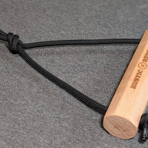 May include: A light-colored wooden handle with the text "RUSTIC REPS-" is connected to a black rope loop. The handle has a hexagonal shape. The rope is knotted at both ends of the handle.