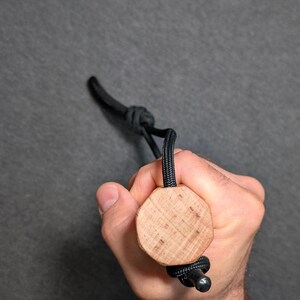 May include: A wooden bottle stopper with a black cord and a metal bead. The stopper is held in a hand against a gray background. The wooden part is a light brown color and has a circular shape.