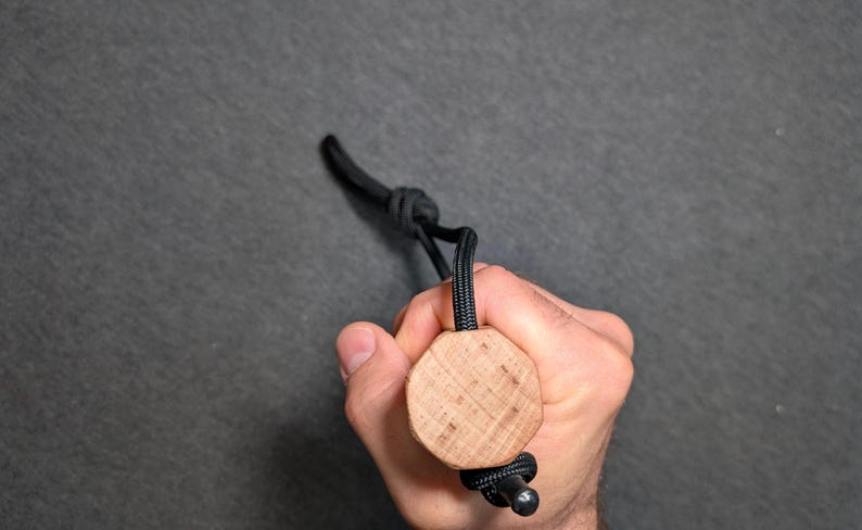 May include: A wooden bottle stopper with a black cord and a metal bead. The stopper is held in a hand against a gray background. The wooden part is a light brown color and has a circular shape.