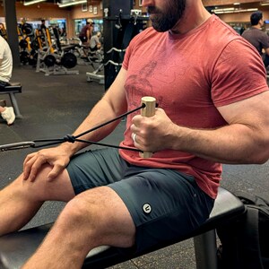 May include: A person using a cable row machine in a gym. The person is wearing a red t-shirt and dark green shorts. The machine has a wooden handle and black cables. Other people and gym equipment are visible in the background.
