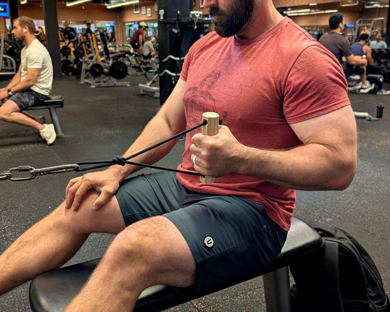 May include: A person using a cable row machine in a gym. The person is wearing a red t-shirt and dark green shorts. The machine has a wooden handle and black cables. Other people and gym equipment are visible in the background.