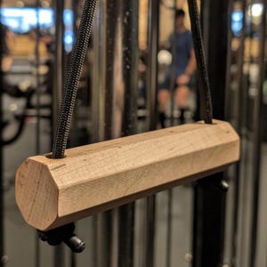 May include: A light brown wooden pull-up bar with a multi-faceted design, suspended by black ropes. The bar is attached to a gym machine. The background is blurred, showing a person in a gym setting.