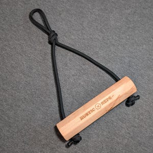 May include: A wooden handle with the text "RUSTIC REPS" is connected to a black cord loop. The handle is light brown and rectangular. The cord is knotted at both ends of the handle.