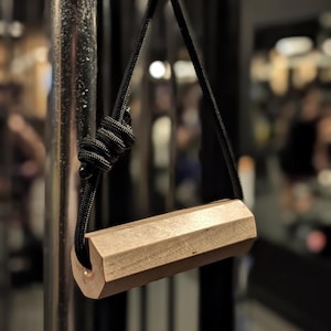 May include: A bronze-colored, hexagonal weightlifting handle with a black rope attached. The handle is suspended from a metal bar, with a close-up view highlighting the texture and design. The rope is knotted at the top, and the background is blurred.