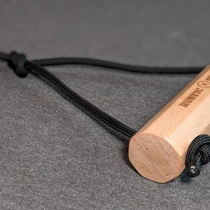 May include: A light-colored wooden handle with the text "RUSTIC REPS" is attached to a black cord. The handle is hexagonal and has a black cord loop at one end. The item is on a gray surface.