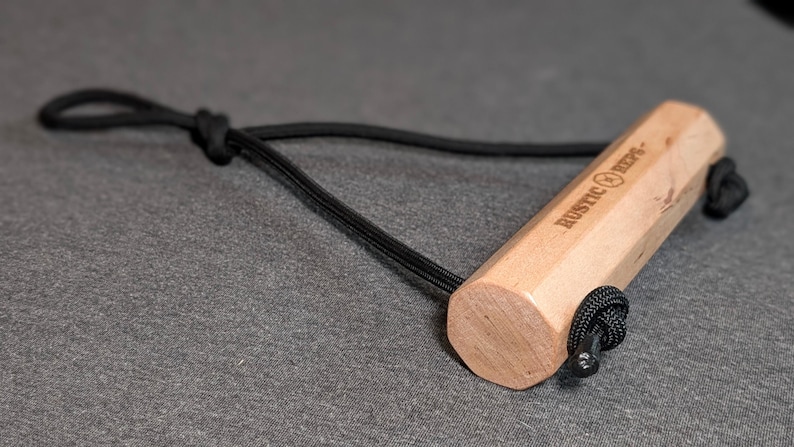 May include: A light-colored wooden handle with the text "RUSTIC REPS" is attached to a black cord. The handle is hexagonal and has a black cord loop at one end. The item is on a gray surface.