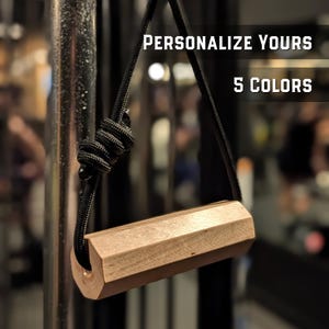 Puede incluir: Un mango de gimnasio de madera clara con forma hexagonal, suspendido por un cordón trenzado negro con un nudo decorativo. El texto "PERSONALIZE YOURS" y "5 COLORS" es visible en la parte superior de la imagen sobre un fondo oscuro.